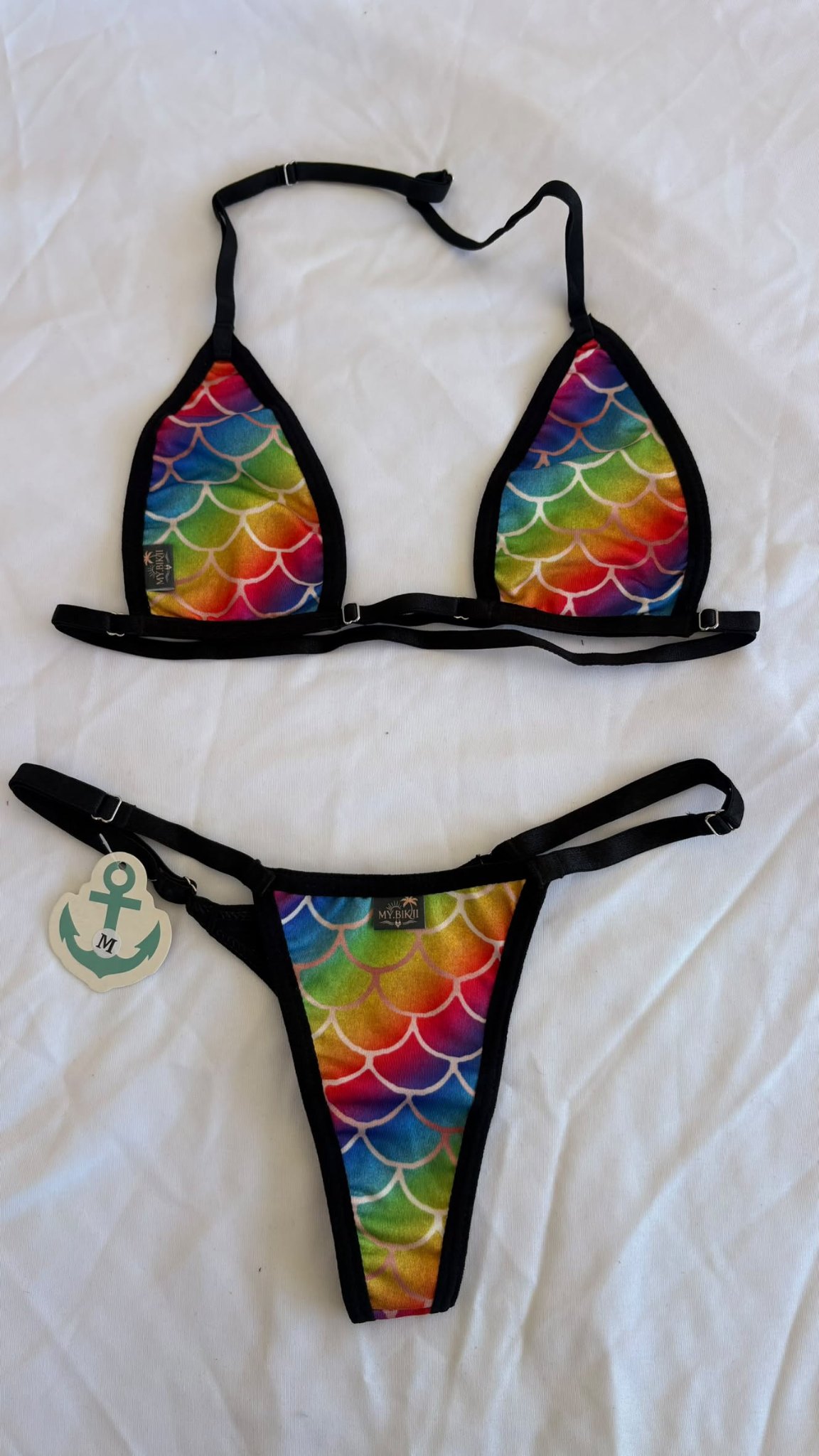 Ensemble bikini triangle à motif écailles multicolores avec liserés noirs, posé à plat sur tissu blanc.